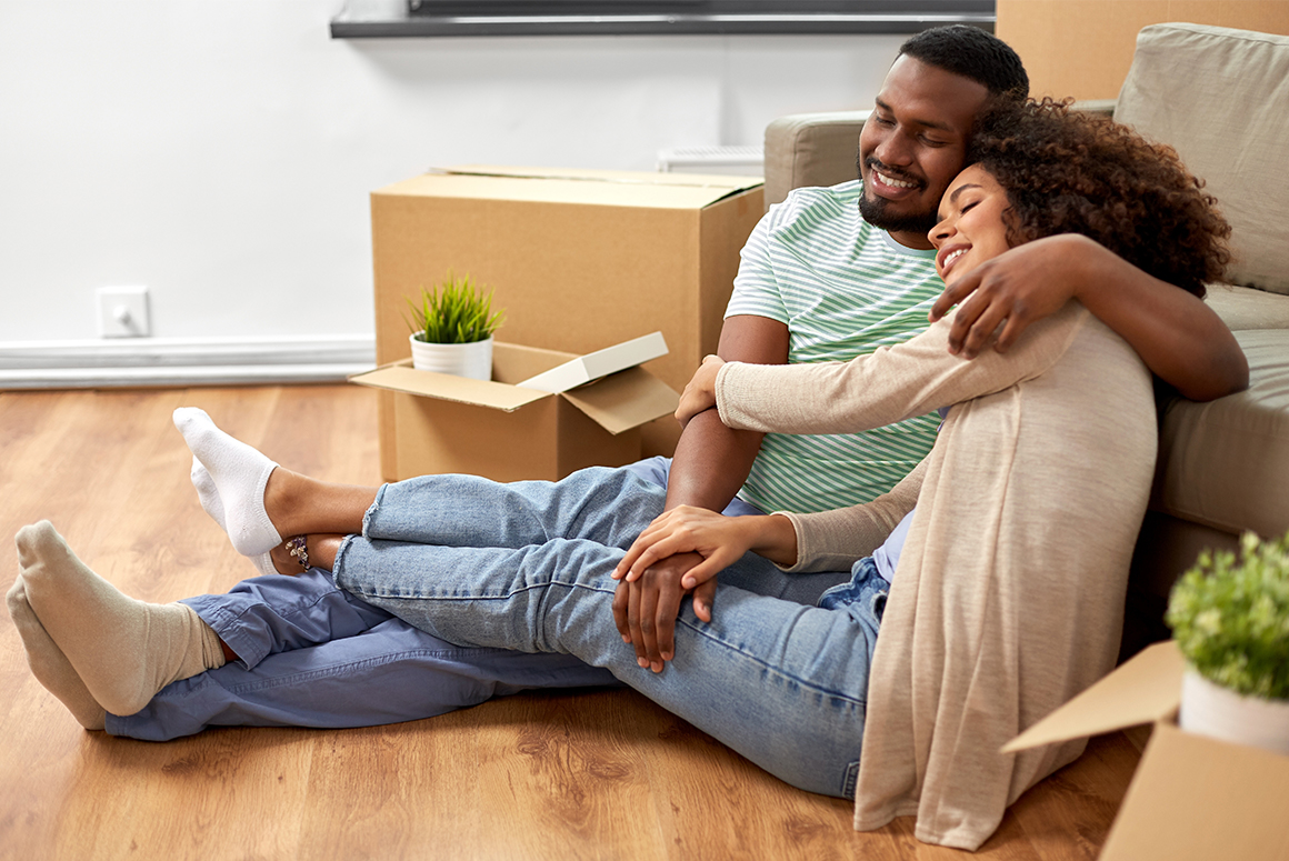 Black couple sitting on the floor of their new home, surrounded by moving boxes