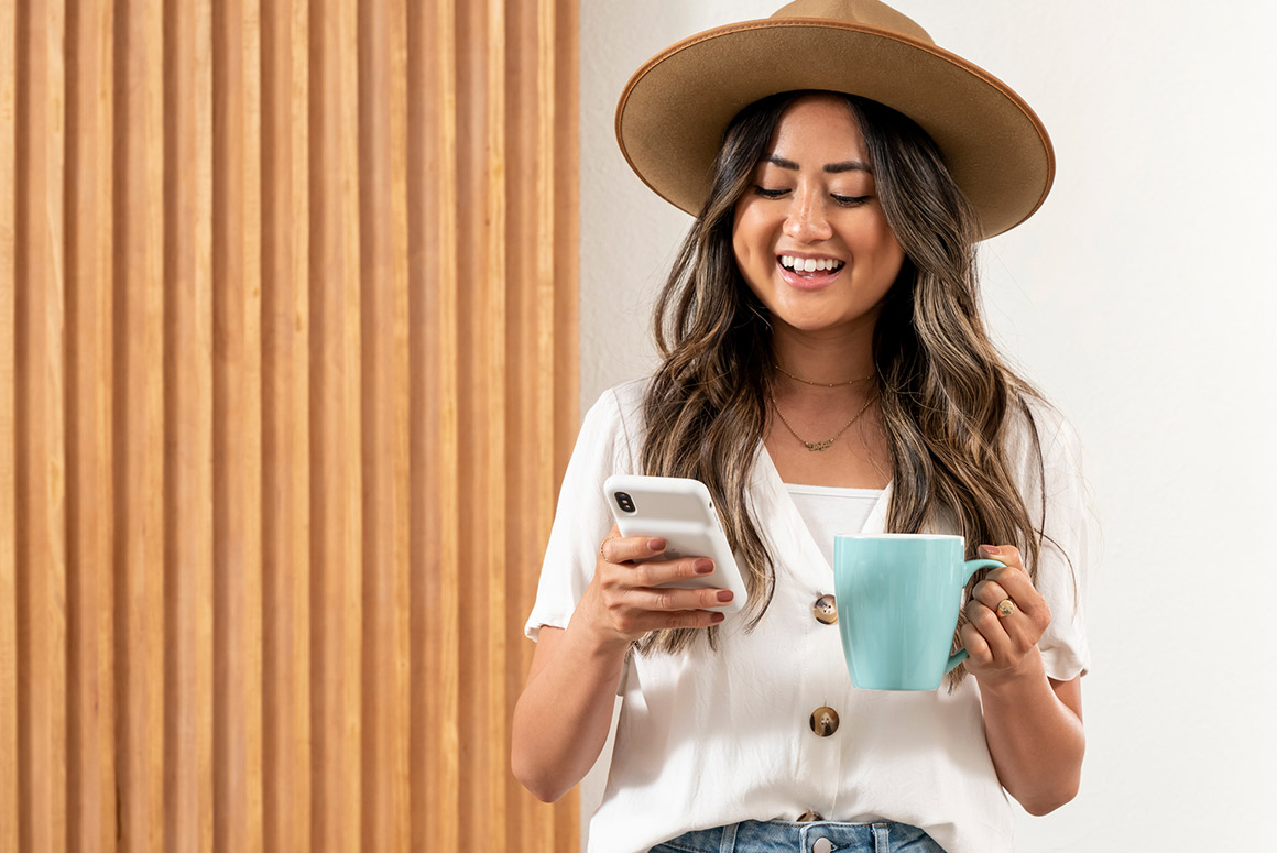 young woman using her smartphone to shop for homes while drinking a cup of coffee