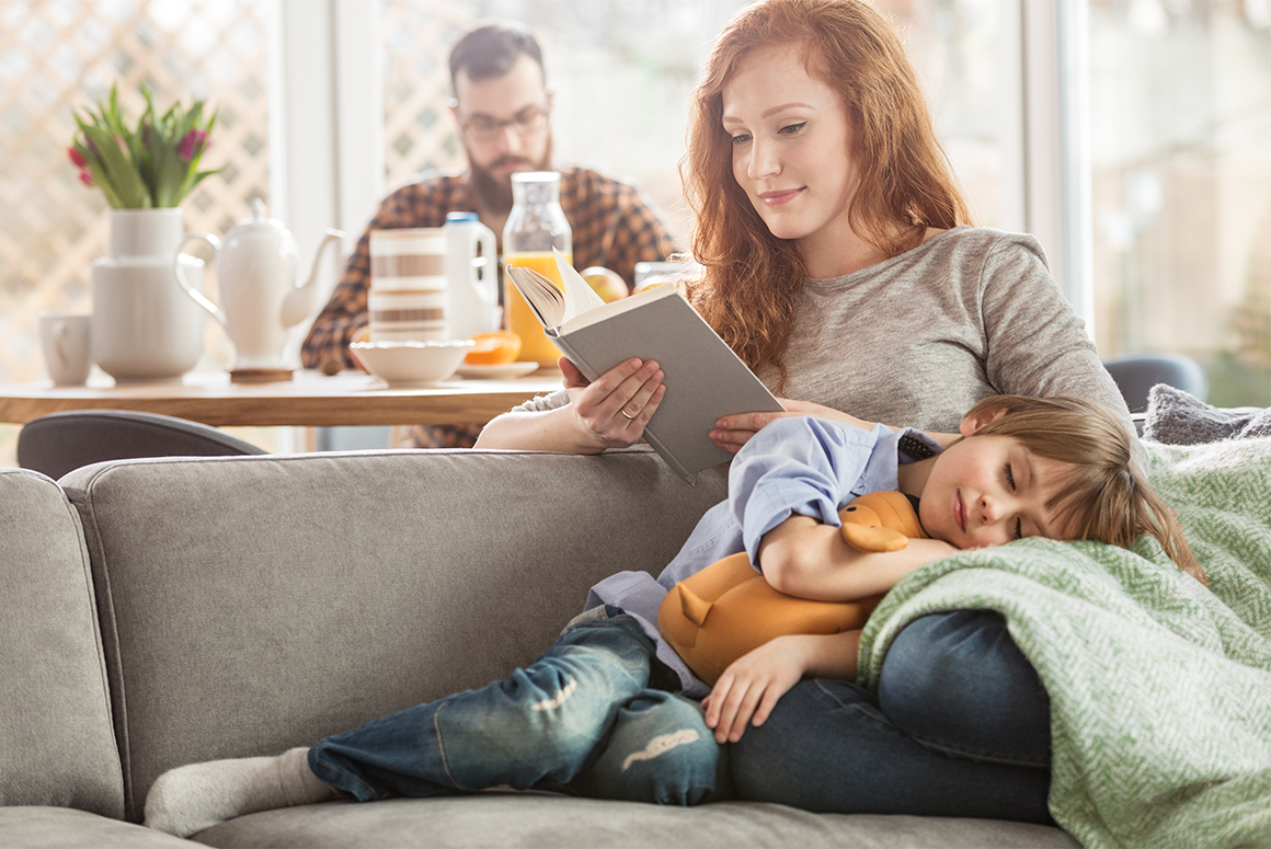 mom and son cuddling on the couch, looking at a tablet. the husband sits at a table behind them.